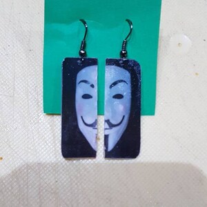 Könnte beinhalten: Ein Paar schwarze Hängeohrringe mit einem weißen und schwarzen Bild der Anonymous-Maske an jedem Ohrring.