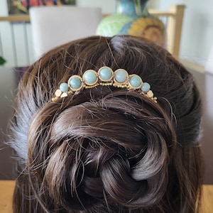 May include: A gold-coloured hair comb with pale blue gemstones is set in dark brown hair. The comb has a curved design with the gemstones evenly spaced across the top, adding a touch of elegance to the hairstyle.