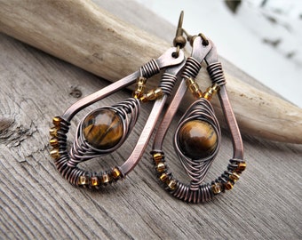 May include: Handmade copper wire earrings with teardrop frames. Each earring features a tiger's eye stone, wrapped in intricate wirework, and accented with small gold-colored beads. The earrings have a rustic, bohemian style.