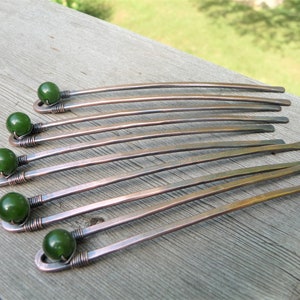 May include: A set of six hair sticks crafted from copper-toned metal, each featuring a smooth, dark green spherical bead secured at the top with coiled wire detailing. The metal shafts are slightly curved and have a flat, hammered finish.