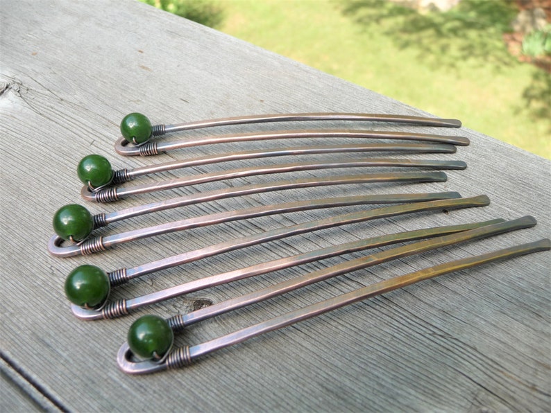 May include: A set of six hair sticks crafted from copper-toned metal, each featuring a smooth, dark green spherical bead secured at the top with coiled wire detailing. The metal shafts are slightly curved and have a flat, hammered finish.