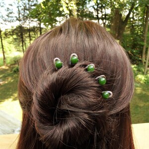 May include: Five silver-toned metal hair pins, each featuring a single round, smooth, dark green bead at the top. The pins are inserted into a brown hair bun, arranged in a curved line following the shape of the hair style to secure it in place.