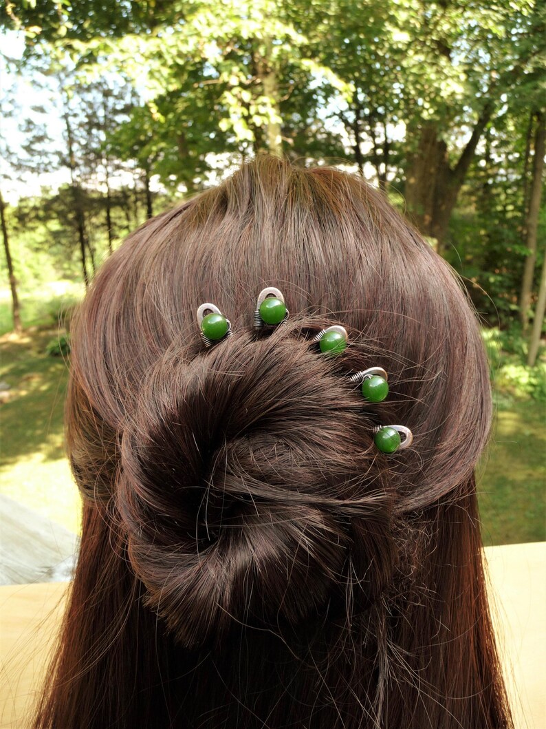 May include: Five silver-toned metal hair pins, each featuring a single round, smooth, dark green bead at the top. The pins are inserted into a brown hair bun, arranged in a curved line following the shape of the hair style to secure it in place.