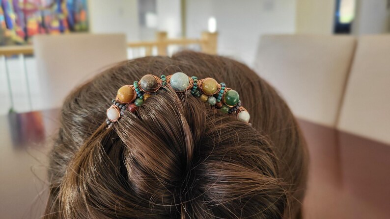 May include: A handcrafted hair accessory featuring a series of colorful, polished gemstones. The stones, in shades of brown, green, and blue, are intricately wired together. This unique hair piece is designed to be worn in the hair.