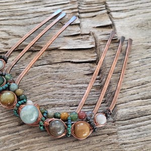 May include: A copper hair comb adorned with various colorful gemstones. The comb features multiple tines and is embellished with green, brown, and cream-colored beads, wrapped in copper wire. The background is a weathered wooden surface.