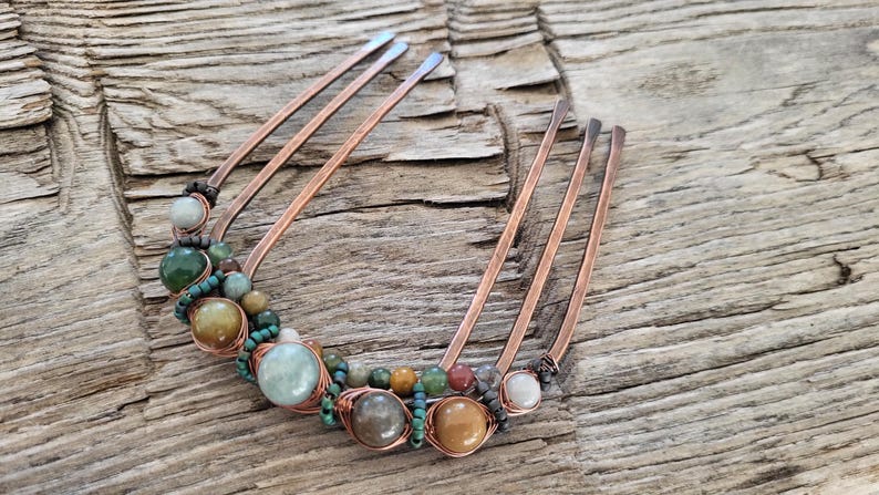 May include: A copper hair comb adorned with various colorful gemstones. The comb features multiple tines and is embellished with green, brown, and cream-colored beads, wrapped in copper wire. The background is a weathered wooden surface.