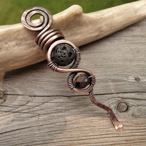 May include: Copper-toned metal pendant with a spiral top and a wavy design. The pendant features two dark, textured stone beads, one round and one faceted, held in place by coiled wire. The pendant is displayed on a wooden surface.
