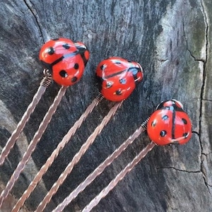 May include: Three ladybird hair pins with red and black painted details. The ladybird toppers are attached to copper-coloured twisted metal stems, set against a weathered wood background.