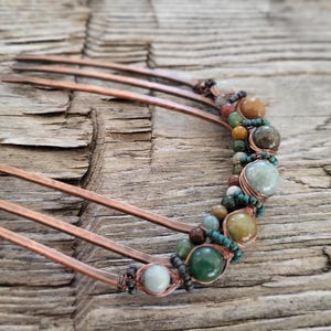 May include: Copper hair comb adorned with a row of colorful beads and stones. The beads are in shades of green, brown, and tan, adding a natural, earthy aesthetic. The comb's tines are made of copper.
