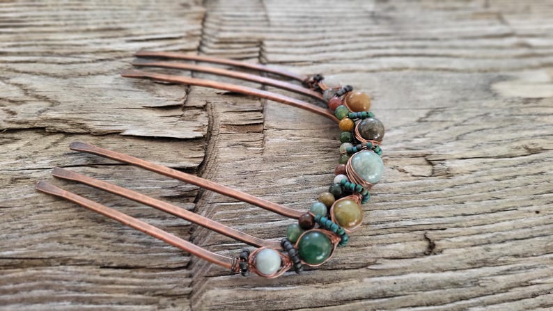 May include: Copper hair comb adorned with a row of colorful beads and stones. The beads are in shades of green, brown, and tan, adding a natural, earthy aesthetic. The comb's tines are made of copper.