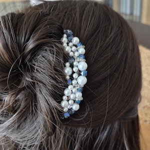 May include: A decorative hair comb with white faux pearls, blue crystals, and clear beads. The comb is set in a dark brown bun hairstyle. This hair accessory is designed for special occasions. The comb is a beautiful addition to any hairstyle.