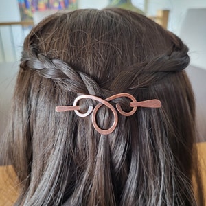 May include: Copper hair pin in an infinity symbol design, set in dark brown hair. The pin is a warm, reddish-brown color and appears to be handmade. The hair is styled in a braid, with the pin securing the hair in place.
