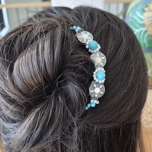 May include: A curved hair accessory featuring a series of silver-toned circular metal discs with intricate floral patterns, interspersed with bright turquoise beads and small white pearls, secured in a dark hair bun for decorative styling.