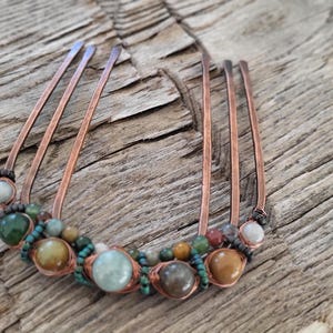 May include: A copper hair comb with five prongs, adorned with a variety of colorful beads. The beads include shades of green, brown, and cream, and are strung together with copper wire. The comb is set against a rustic wooden background.