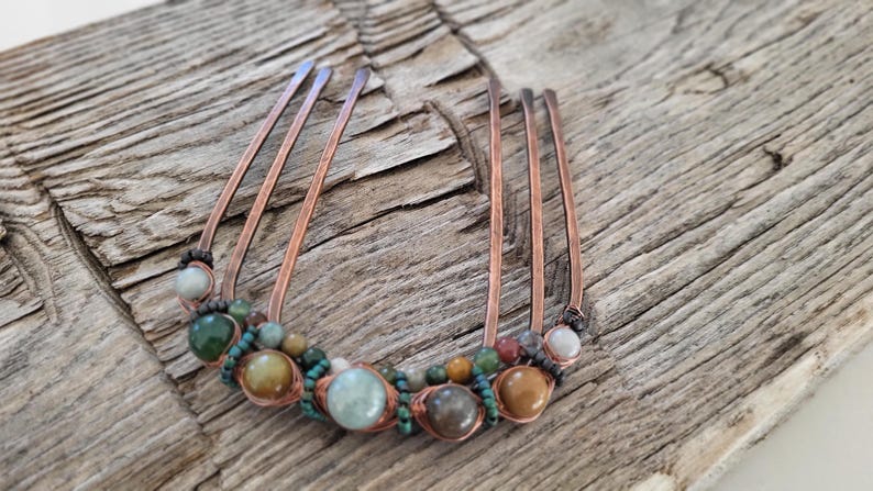 May include: A copper hair comb with five prongs, adorned with a variety of colorful beads. The beads include shades of green, brown, and cream, and are strung together with copper wire. The comb is set against a rustic wooden background.