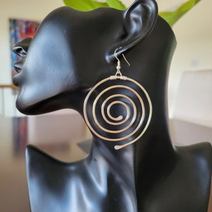 May include: Silver spiral earrings on a black mannequin head. The earrings have a large, open spiral design, with a hook for pierced ears. The earrings are a bright silver colour and appear to be made of metal.