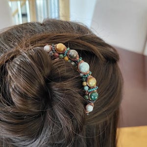 May include: A decorative hair comb featuring a curved design adorned with various colorful gemstones. The gemstones include shades of brown, green, and white, set against a copper-colored wire. The comb is inserted into a bun.