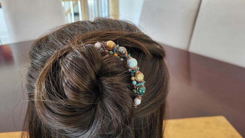 May include: A decorative hair comb featuring a curved design adorned with various colorful gemstones. The gemstones include shades of brown, green, and white, set against a copper-colored wire. The comb is inserted into a bun.