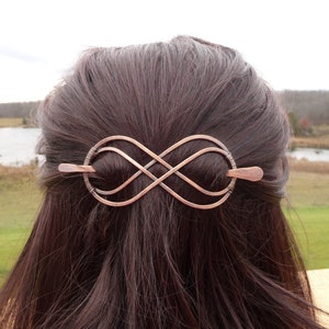 May include: Copper-colored hair barrette in an infinity symbol design. The barrette is made of bent metal wire and features a long pin for securing it in the hair. The barrette is approximately 4 inches long.