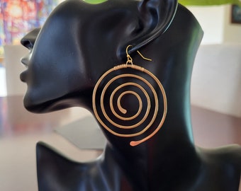 May include: Gold-colored spiral earrings with a hook closure. The earrings are displayed on a black mannequin head. The spiral design is a prominent feature, with the metal wire forming concentric circles. The earrings are a statement piece.