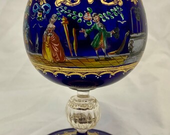 Op de afbeelding: Een kobaltblauw glazen kelk met een gouden en wit geschilderd ontwerp. De kelk toont een scène van mensen in een landschap. De kelk heeft een heldere glassteel en een ronde, heldere glazen basis.