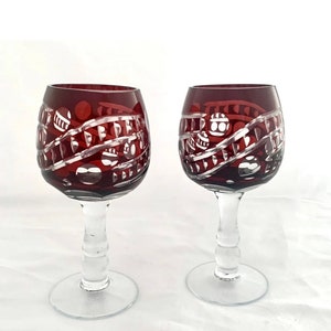 May include: Two clear glass wine glasses with a burgundy red stem and bowl. The bowl has a cut glass design with a pattern of circles and lines.