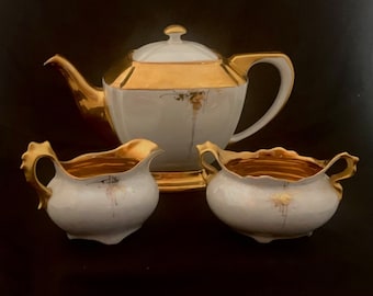 Puede incluir: Un juego de té de porcelana blanca con detalles dorados. La tetera, la jarra de crema y el azucarero tienen un delicado diseño floral con ribetes dorados.