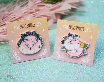 May include: Two pink enamel pins featuring a curled up snake with green leaves and flowers. The pins are on a pink background with the text "Shiny Snakes" on each card.