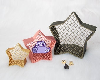 May include: Three star-shaped decorative objects made of a wire mesh material. The largest star is dark brown, the middle star is pink, and the smallest star is gold. The middle star has a purple cartoon raccoon with a trash can on it. There are two gold pin backs on the white surface.