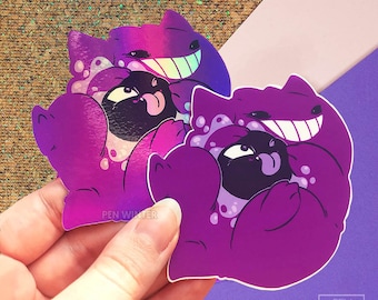 May include: Two purple and black holographic stickers of a cartoon cat with a wide grin and a smaller cat with a tongue sticking out. The text "PEN WINTER" is visible on the sticker.