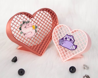 May include: Two heart-shaped pin holders, one pink and one copper, with a grid pattern. The pink holder has a purple raccoon pin, and the copper holder has a pink snake pin.