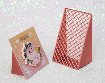 May include: A pink and white card with the text "Shiny Snakes" and a cartoon illustration of a pink snake with yellow accents. The card is propped up on a pink triangular display stand with a grid pattern.