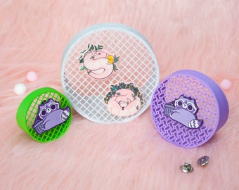 May include: Three enamel pins featuring a raccoon, a snake, and a sleeping cat. The pins are displayed on a white, green, and purple gridded background. The pins are all purple and feature a white outline. The raccoon pin features a trash can. The snake pin features a green plant. The cat pin features a pink flower.