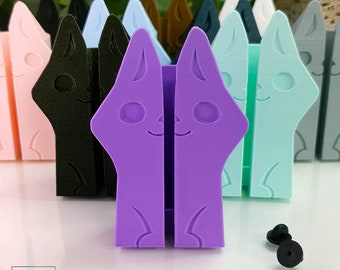 May include: A set of 10 colorful cat-shaped pen holders, each with a different color. The pen holders are made of plastic and have a cute cartoon cat face on the front. The pen holders are arranged in a row, with the purple one in the center. There are two black pin backs on the white surface below the pen holders.