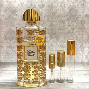 May include: A clear perfume bottle with gold floral detailing and a gold cap, labeled "Sublime Vanille." Accompanied by three smaller gold-capped spray bottles. The perfume bottle has a gold emblem and the text "CREED 1760".