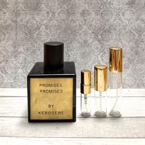 May include: A black rectangular perfume bottle with a gold label that reads "PROMISES, PROMISES BY KEROSENE." Next to it are three clear glass spray bottles with gold caps. The bottles are of varying sizes.