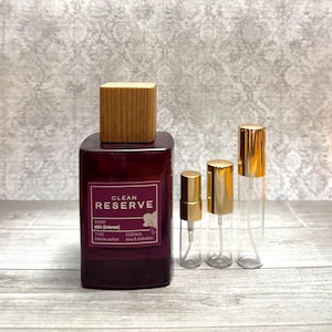 May include: A rectangular, dark red perfume bottle with a wooden cap, labeled "Clean Reserve." Three smaller, clear spray bottles with gold caps are next to it. The perfume bottle has a white label with the scent name and description.