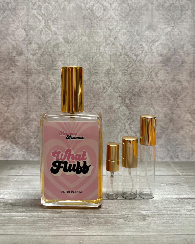 May include: A clear glass perfume bottle with a gold cap and a pink label that reads "What the Flubb" in black script. Two smaller spray bottles and a travel-sized bottle with gold caps are next to the perfume bottle. The words "Aromas" and "Eau de Parfum" are also on the label.