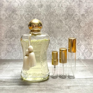 May include: A clear glass perfume bottle with a gold cap and a white tassel, alongside three clear glass spray bottles with gold caps. The perfume bottle is filled with a light yellow liquid.