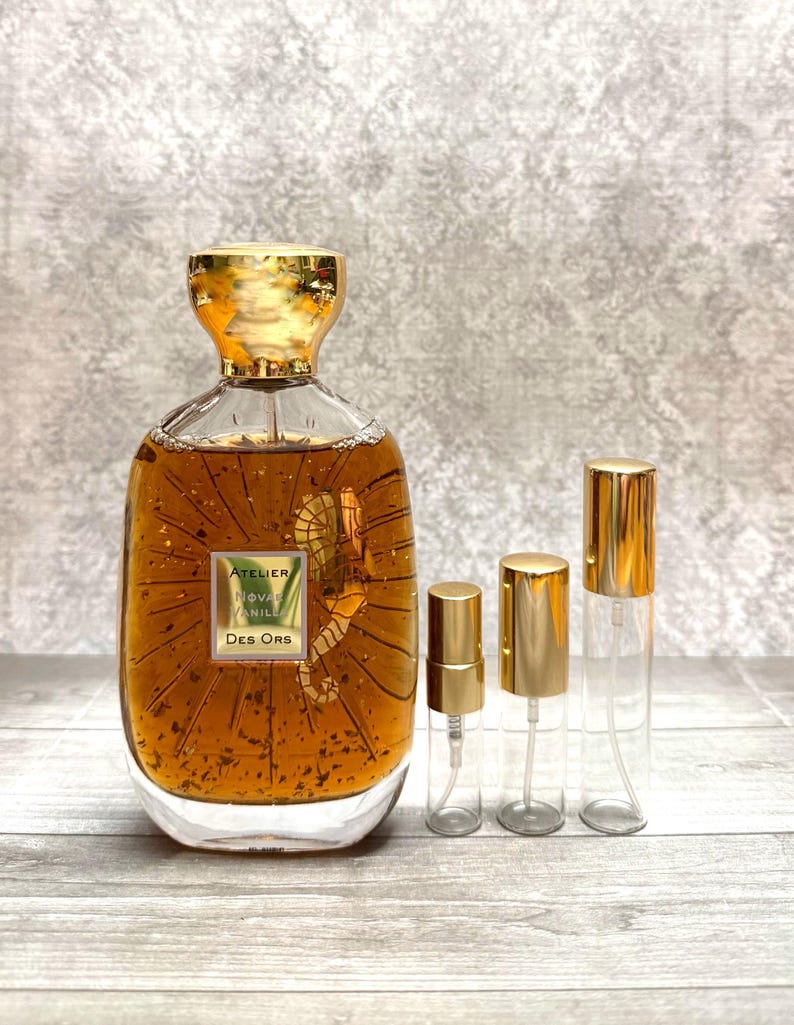 May include: A perfume bottle and three smaller spray bottles. The large bottle is filled with a golden liquid and has a gold cap. The label reads "ATELIER NOVA VANILLE Des Ors". The smaller bottles have gold caps.