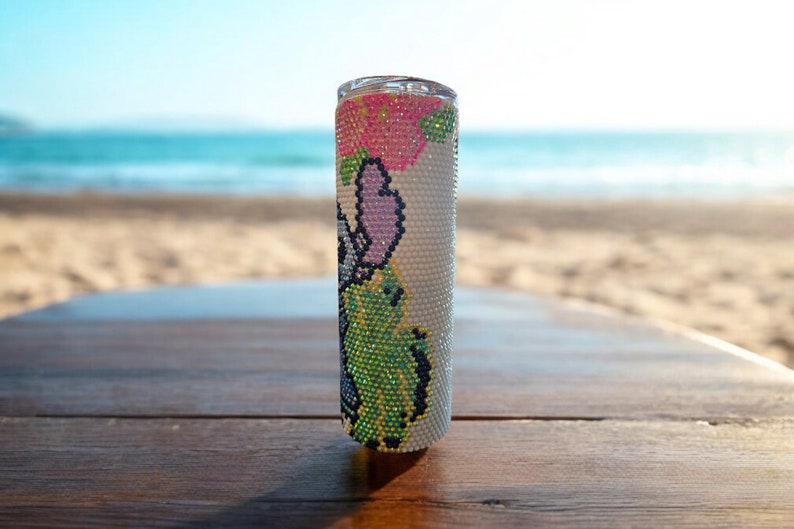 Op de afbeelding: Een witte tumbler met een kleurrijk bloemenontwerp met roze, groene en paarse bloemen. De tumbler is gemaakt van roestvrij staal en heeft een doorzichtig deksel.