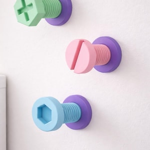 May include: Three colourful wall hooks designed as oversized screws. The hooks have green, pink, and blue screw heads with a purple base. The screws are mounted on a white wall, providing a playful and functional design element.