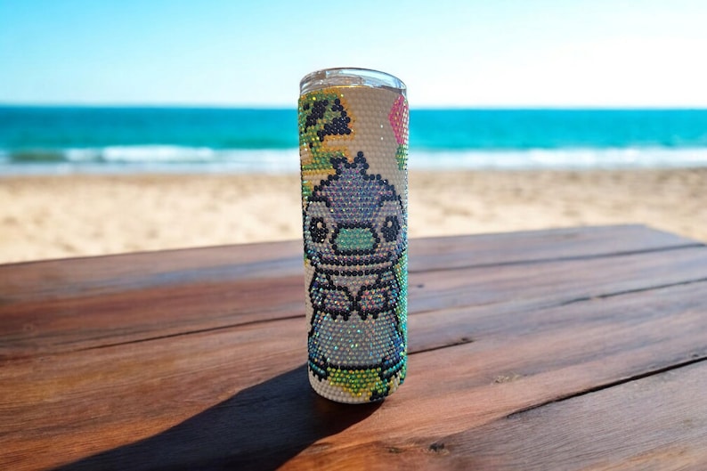 Op de afbeelding: Een witte roestvrijstalen tumbler met een kleurrijk ontwerp met een cartoonfiguur. Het figuur is blauw, groen en roze en heeft grote ogen. De tumbler staat op een houten tafel.