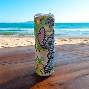 Op de afbeelding: Een witte tumbler met een kleurrijk cartoon-personage ontwerp van Stitch uit Lilo & Stitch. De tumbler staat op een houten oppervlak met een strand op de achtergrond.