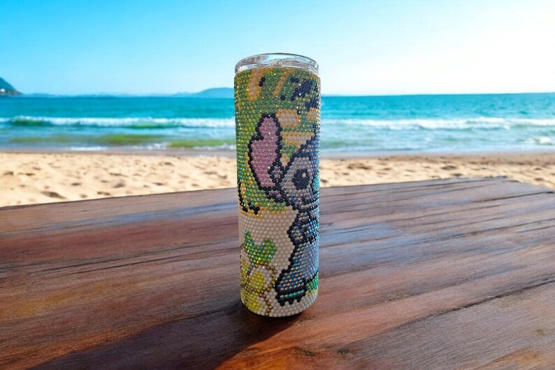 Op de afbeelding: Een witte tumbler met een kleurrijk cartoon-personage ontwerp van Stitch uit Lilo & Stitch. De tumbler staat op een houten oppervlak met een strand op de achtergrond.