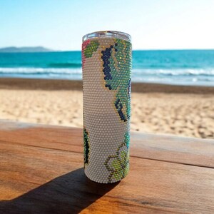 Op de afbeelding: Een witte tumbler met een groen, blauw en roze bloemenontwerp. De tumbler staat op een houten oppervlak met een strand op de achtergrond.