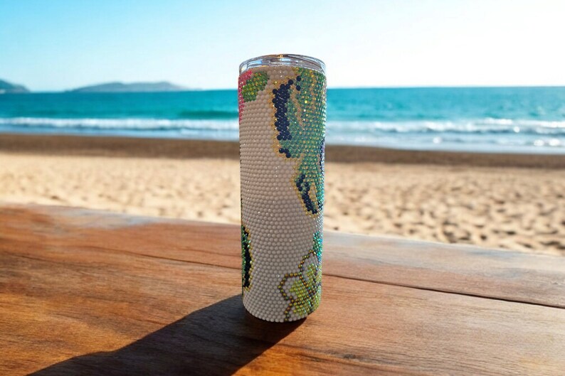 Op de afbeelding: Een witte tumbler met een groen, blauw en roze bloemenontwerp. De tumbler staat op een houten oppervlak met een strand op de achtergrond.