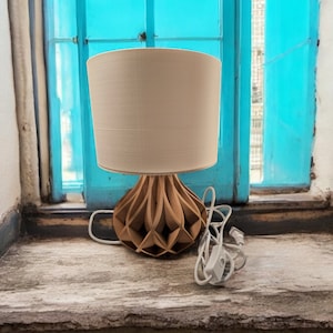 May include: A wooden table lamp with a white fabric shade. The lamp is sitting on a windowsill with a blue window behind it.