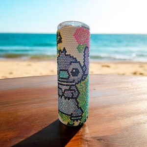 Op de afbeelding: Een witte roestvrijstalen tumbler met een kleurrijk ontwerp van strass steentjes met een cartoonfiguur. Het ontwerp bevat een blauw figuur met roze oren en een roze bloem.
