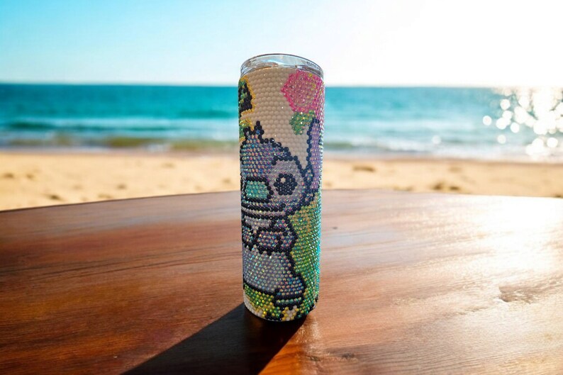 Op de afbeelding: Een witte roestvrijstalen tumbler met een kleurrijk ontwerp van strass steentjes met een cartoonfiguur. Het ontwerp bevat een blauw figuur met roze oren en een roze bloem.